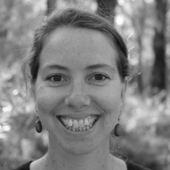 A woman smiling outdoors in a black-and-white photo, surrounded by softly blurred trees and sunlight.
